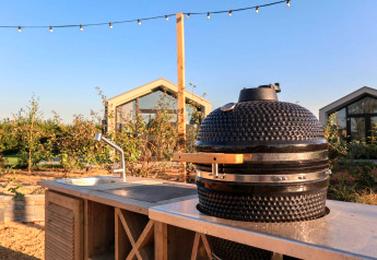 Udendørs køkken med keramisk grill og vask på en glampingplads, med drivhuse i baggrunden under blå himmel.