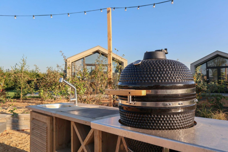 Outdoor kitchen with ceramic grill and sink at a glamping site, greenhouses visible in the sunny background.