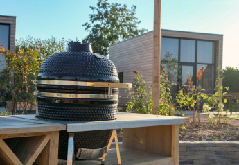 Outdoor glamping scene with a ceramic barbecue grill on a counter and a modern wooden cabin in the background.
