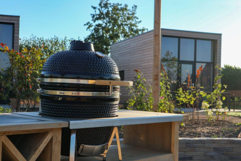 Escena de glamping al aire libre con barbacoa de cerámica sobre una mesa y cabaña moderna al fondo.