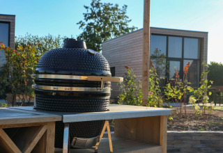 Escena de glamping al aire libre con barbacoa de cerámica sobre una mesa y cabaña moderna al fondo.