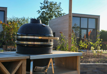 Escena de glamping al aire libre con barbacoa de cerámica sobre una mesa y cabaña moderna al fondo.