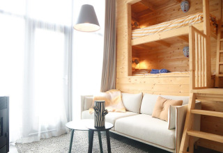 Licht interieur met houten stapelbedden, comfortabele bank, tafel en grote ramen in Tiny House 4, Nederland.