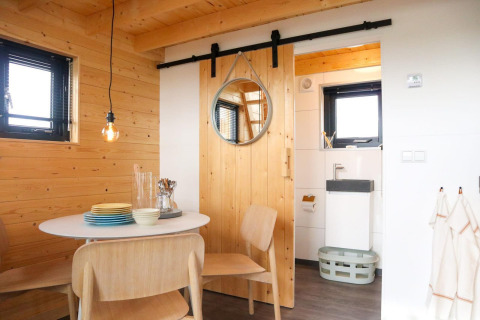 Moderne, compacte leefruimte met eettafel, houten stoelen en zicht op badkamer in Tiny House 4, Nederland.