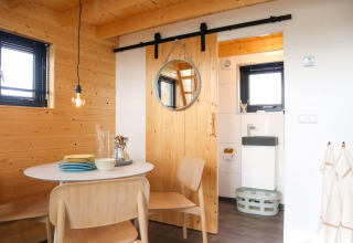 Moderne, compacte leefruimte met eettafel, houten stoelen en zicht op badkamer in Tiny House 4, Nederland.