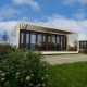 Moderne lodge Modus 4 med glasfacade og åben ramme, omgivet af frodige haver under blå himmel.