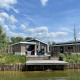 Moderne Hackfort 6 hytter ved søen med træterrasse, grønne omgivelser og blå himmel med skyer.
