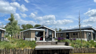 Modernos alojamientos Hackfort 6 junto al lago, con terraza de madera, césped y cielo azul con nubes.