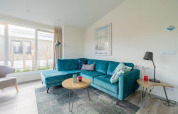 Lichte woonkamer met turquoise hoekzetel, moderne tafeltjes en grote ramen in Hackfort Waterfront 4.