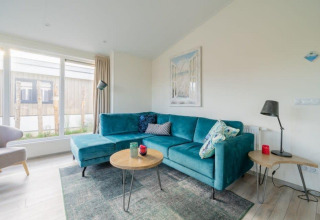 Lichte woonkamer met turquoise hoekzetel, moderne tafeltjes en grote ramen in Hackfort Waterfront 4.