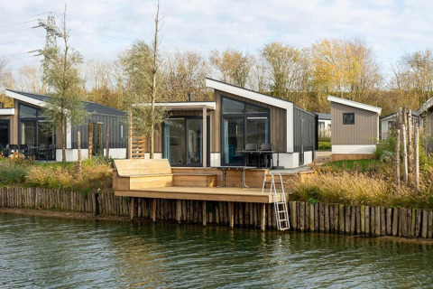 Moderne Hackfort Waterfront 8 Lodge mit Terrasse am Wasser, umgeben von Bäumen und Natur.