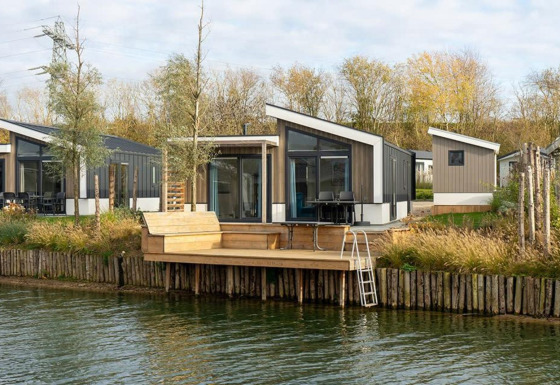Moderne Hackfort Waterfront 8 Lodge mit Terrasse am Wasser, umgeben von Bäumen und Natur.