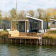 Moderne lodge Hackfort Waterfront 8 med stort vinduesparti ved vandkanten, omgivet af træer og natur.