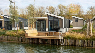 Moderne Hackfort Waterfront 8 Lodge mit Terrasse am Wasser, umgeben von Bäumen und Natur.