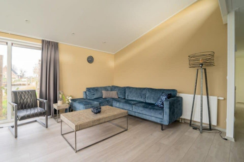 Sala moderna en la cabaña Hackfort Waterfront 8 con sofá azul, pared beige y grandes ventanales en Países Bajos.