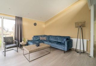 Sala moderna en la cabaña Hackfort Waterfront 8 con sofá azul, pared beige y grandes ventanales en Países Bajos.