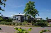 Moderne glampingaccommodatie met grote ramen, terras en groene beplanting onder een blauwe lucht.
