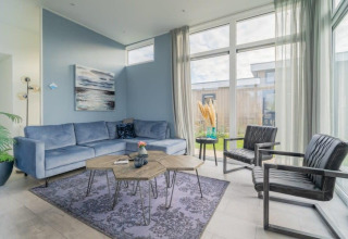 Stijlvolle loungehoek in een lodge met blauwe zetel, zwarte stoelen, grote ramen en moderne inrichting.