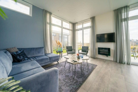 Moderne lodge-woonkamer met grote ramen, blauwe hoekbank, wand-tv en stijlvolle inrichting.
