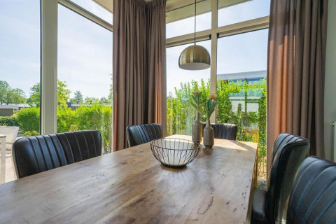 Luminoso comedor en una cabaña con sillas negras, mesa de madera, grandes ventanales y vista al jardín.