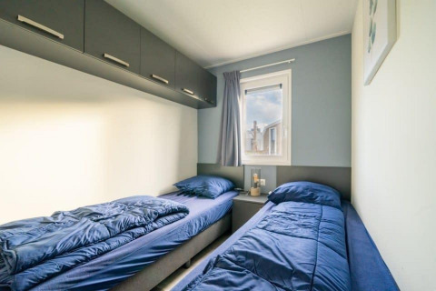 Chambre de lodge moderne avec deux lits simples, literie bleue, placards en hauteur et fenêtre avec rideau.