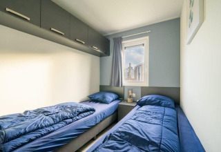 Chambre de lodge moderne avec deux lits simples, literie bleue, placards en hauteur et fenêtre avec rideau.