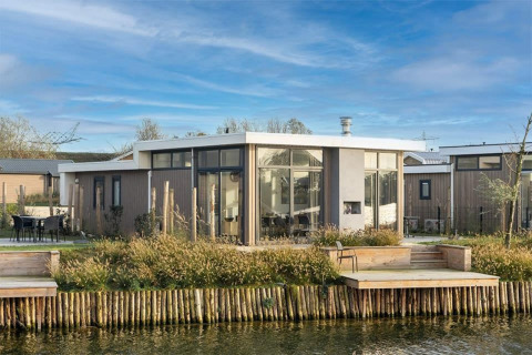Moderne Pavilion Waterfront 8 lodge met grote ramen en terras aan het water, omringd door groen landschap.