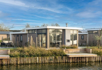 Moderne Pavilion Waterfront 8 lodge med store vinduer og terrasse ved vandet, blå himmel og grønne omgivelser.
