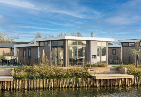 Moderne Pavilion Waterfront 8 lodge met grote ramen en terras aan het water, omringd door groen landschap.