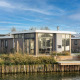 Moderne Pavilion Waterfront 8 lodge med store vinduer og terrasse ved vandet, blå himmel og grønne omgivelser.