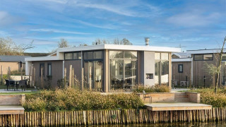 Moderne Pavilion Waterfront 8 lodge med store vinduer og terrasse ved vandet, blå himmel og grønne omgivelser.