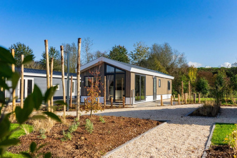 Lodge moderne aux grandes baies vitrées, entouré d’arbres et de sentiers paysagers sous un ciel bleu.
