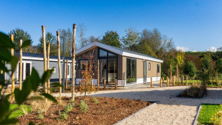Lodge moderne aux grandes baies vitrées, entouré d’arbres et de sentiers paysagers sous un ciel bleu.