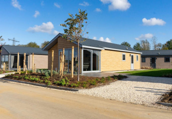 Moderne lodge met lichte houten bekleding, tuin en grindoprit onder een heldere blauwe hemel met wolken.