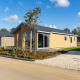 Moderne lodge med lyse træfacader, haveanlæg og grusindkørsel under en klar blå himmel med skyer.