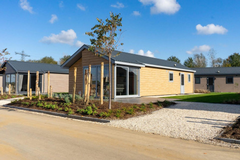 Lodge moderne en bois clair avec jardin paysager et allée de gravier sous un ciel bleu et ensoleillé.
