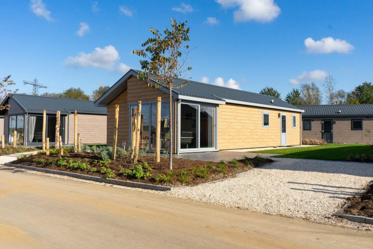 Moderna cabaña de madera clara, jardín cuidado y entrada de grava bajo un cielo azul y soleado.