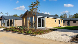 Lodge moderne en bois clair avec jardin paysager et allée de gravier sous un ciel bleu et ensoleillé.