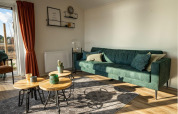 Moderne lodge woonkamer met groene zetel, ronde houten tafels, rode gordijnen en veel natuurlijk licht.