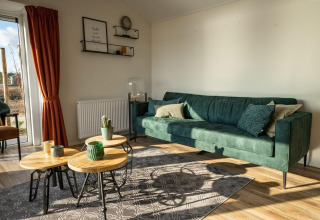 Modernes Lodge-Wohnzimmer mit grünem Sofa, runden Holztischen, roten Vorhängen und viel Tageslicht.