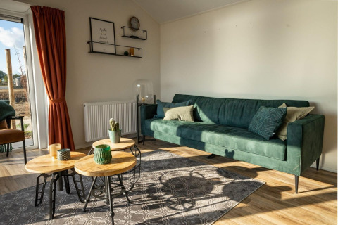 Salón moderno de lodge con sofá verde, mesas redondas de madera, cortinas rojas y abundante luz natural.