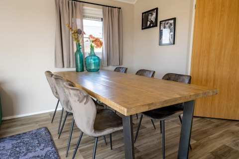 Spisebord i træ med seks stole, grønne vaser og blomster, ved siden af vindue med lyse gardiner i en lodge.