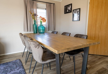 Wooden dining table with six chairs, green vases and flowers, near window with curtains inside a lodge.