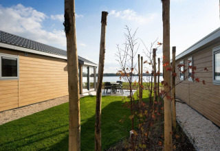 Udsigt mellem to træbeklædte hytter ved en sø, med græsplæne og træer under blå himmel.