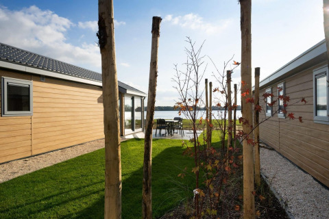 Udsigt mellem to træbeklædte hytter ved en sø, med græsplæne og træer under blå himmel.