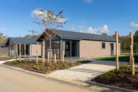 Moderne lodge med træbeklædning og store vinduer, omgivet af have og blå himmel på en solrig dag.