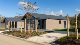Lodge moderne en bois avec de grandes fenêtres, entouré de jardins et sous un ciel bleu lumineux.