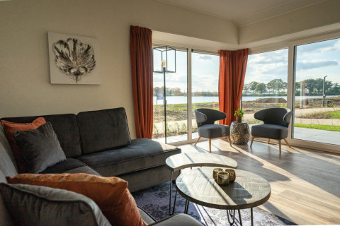 Helles Lodge-Wohnzimmer mit großen Fenstern, Blick auf einen See, grauem Sofa und orangen Vorhängen.