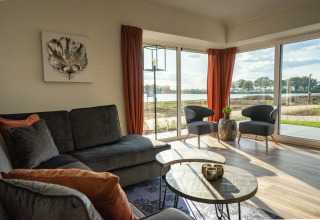 Helles Lodge-Wohnzimmer mit großen Fenstern, Blick auf einen See, grauem Sofa und orangen Vorhängen.