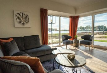 Lyst og moderne lodge-stue med store vinduer, udsigt til sø, grå sofa og orange gardiner.